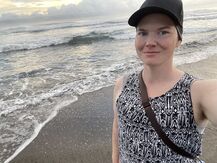JJ Bunch is standing on a beach with wet sand and clouds covering the sun behind their shoulder. Waves are visible with whitecaps and are grey blue. JJ is wearing a black hat and is looking directly at the camera and smiling with their mouth closed. They have a black and white patterned tank top on with a black over-the-shoulder bag strap visible across their body.