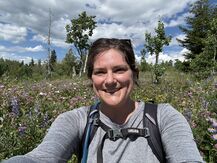 Image of Amanda Kieval sitting in a wildflower field and smiling directly at the camera. The sky behind her is blue and dappled with fluffy white and gray clouds; there are trees on the horizon; and pink, purple, blue, and yellow wildflowers are visible in a green field. Amanda has brown hair pulled back with the exception of side-swept bangs, has a gold septum ring, and is smiling with their teeth visible. She is wearing a long-sleeve heathered grey shirt with Camelbak backpack straps with chest strap visible on top.