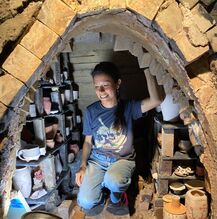 Image of Paola Evangelista crouching and smiling inside of a kiln with ceramic work stacked to the side. Paola's hair is dark and pulled back into a ponytail which is laying over her shoulder. A dangling earring is visible next to her ponytail. Her head is tilted down and her gaze is ahead of her and to the side. She is wearing a dark blue heathered t-shirt with a white skull design on it and light-washed denim jeans.