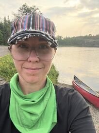 Image of Mel Timpano standing and smiling next to a tree-lined and calm lake with a red canoe on the shore. The sun is behind their shoulder and is obscured by clouds. Mel is looking directly at the camera wearing a multi-colored textile cap with frayed brim, clear round-rimmed glasses, and a green scarf around her neck over a black long-sleeve top.
